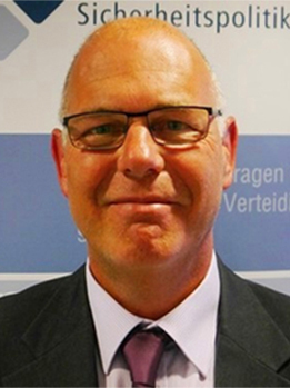 Nahaufnahme eines lächelnden, glatzköpfigen Mannes mit Brille, Anzug und Krawatte, im Hintergrund teilweise 'Sicherheitspolitik'.
