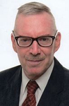 Kopfaufnahme eines älteren Mannes mit grauem Haar, Brille, Schnurrbart, Anzug und Krawatte, der leicht lächelnd in die Kamera blickt.