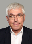 Porträtaufnahme eines lächelnden Mannes mittleren Alters mit grauem Haar, Brille und dunklem Anzug vor grauem Hintergrund.