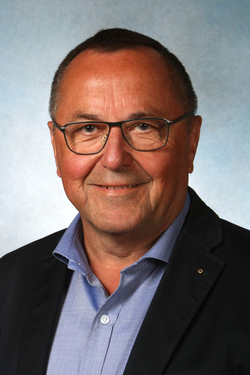 Lächelnder Mann mit Brille, blauem Hemd und dunklem Sakko vor blauem Hintergrund.