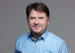 Ein lächelnder Mann mit kurzen dunklen Haaren in einem hellblauen Hemd, der direkt in die Kamera blickt, vor grauem Hintergrund.
