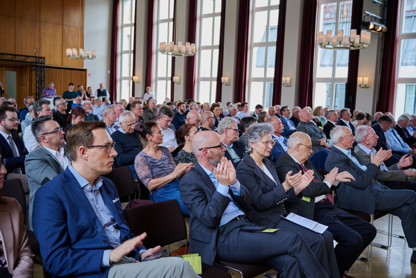 Zahlreiche Personen in einem hellen Konferenzsaal applaudieren. Der Raum hat hohe Fenster und Kronleuchter.