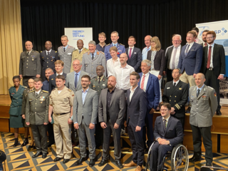 Diverse Gruppe von Militärangehörigen und Zivilisten, einige in Uniform, andere in Anzügen, bei einer Veranstaltung.