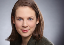 Porträt einer lächelnden Frau mit hellbraunem Haar, blauen Augen und Ohrsteckern, die eine Tweedjacke vor grauem Hintergrund trägt.