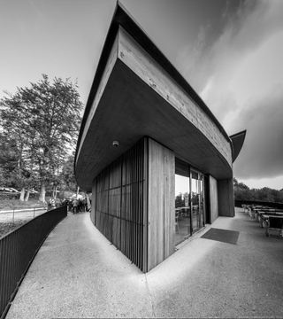 Monochrome Ansicht eines modernen Gebäudes mit geschwungenem Betondach, Holzverkleidung und Glastüren, neben einem Weg mit Menschen.
