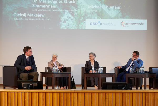 Vier Personen, zwei Männer, zwei Frauen, sitzen auf einer Bühne vor einer Leinwand mit Namen und Logos einer Podiumsdiskussion.