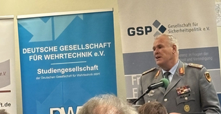 Neun Personen, darunter zwei Frauen und sieben Männer, stehen in einer Reihe vor Veranstaltungsbannern. Eine Frau hält einen Strauß.