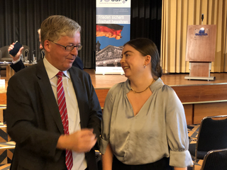 Ein lächelnder Mann im Anzug unterhält sich mit einer lächelnden Frau in einer Bluse vor einem Banner zur deutschen Sicherheitspolitik.