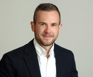 Porträt eines lächelnden jungen Mannes mit Bart, dunklem Blazer und weißem Hemd vor hellem Hintergrund.