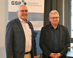 Zwei lächelnde Männer in Business-Casual-Kleidung, einer mit Schnurrbart, posieren vor einem 'GSP Gesellschaft für Sicherheitspolitik e.V.' Banner.