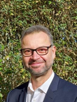 Ein lächelnder Mann mit kurzem grauem Haar, Brille und Bart, Anzugjacke und weißem Hemd, vor grünem Laub.