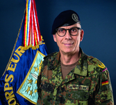 Lächelnder deutscher Soldat Brinkmann in Flecktarnuniform und Barett, mit einer 'CORPS EUR'-Flagge im Hintergrund.